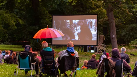 Zuschauer*innen beim Jugendforum Sommerkino, im Vordergrund eine Person mit buntem Regenschirm, im Hintergrund die Leinwand